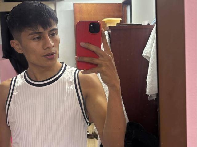 Kyller69, un giovane asiatico con i capelli neri corti, che indossa una canotta bianca a coste, tiene in mano un telefono rosso e si sta scattando una foto allo specchio. selfie, contorno visibile del pene sotto la canotta, espressione seria.