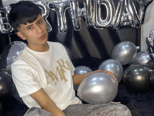 Kyller69, een jonge man met kort zwart haar, die een witte "A&M"-shirt draagt, zilveren ballonnen vasthoudt en zit, verjaardag. Achtergrond met "HAPPY" ballonnen, sexy, ontspannen, nonchalante pose, lichte grijns..