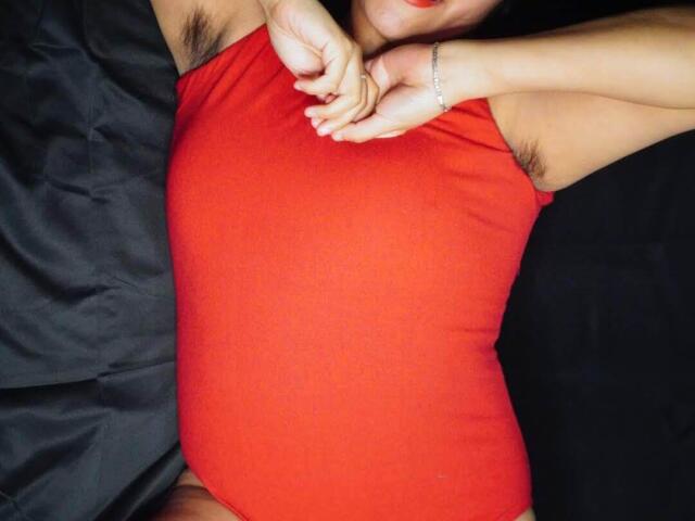 A close-up photo of HairyDuchess lying on a black surface. She wears a tight, bright red one-piece, her arms raised above her head. Her armpits are visible with dark hair. Her left wrist has a silver bracelet. Her red lips contrast with her fair skin. The image focuses on the red one-piece, with the black background providing minimal distraction. The texture of the one-piece is smooth, while the background has a slightly wrinkled, shiny texture.