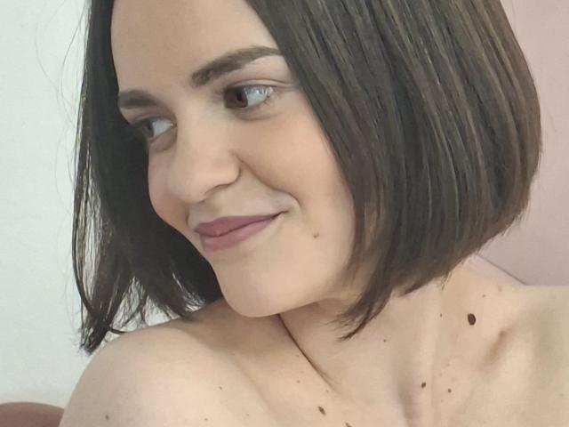 Close-up van BelleWhiteX, een vrouw met een lichte huid en een kort zwart bob kapsel, die subtiel glimlacht. Ze is naakt van boven, en je ziet sproeten en moedervlekken op haar borst. Haar blik is iets naar beneden gericht, haar lippen zijn gesloten..