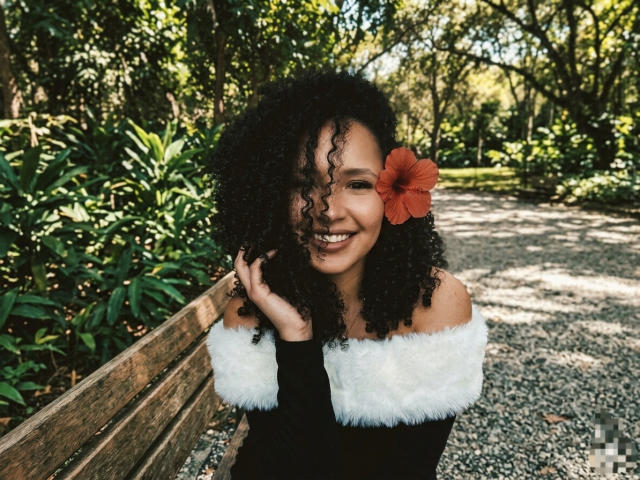 AbvrilDubois with curly black hair and an orange hibiscus flower, smiling, wearing a black top with white fur trim, sitting on a wooden bench in a sunlit garden. Fucking cute.