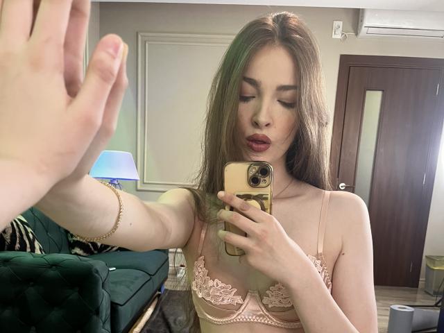 ScarletShow, capelli castani lunghi, reggiseno di pizzo rosa, che si scatta un selfie allo specchio con un telefono dorato, con la mano sollevata di fronte, leggermente. bocca aperta, illuminazione soffusa, ambiente interno, sottile tensione sessuale.