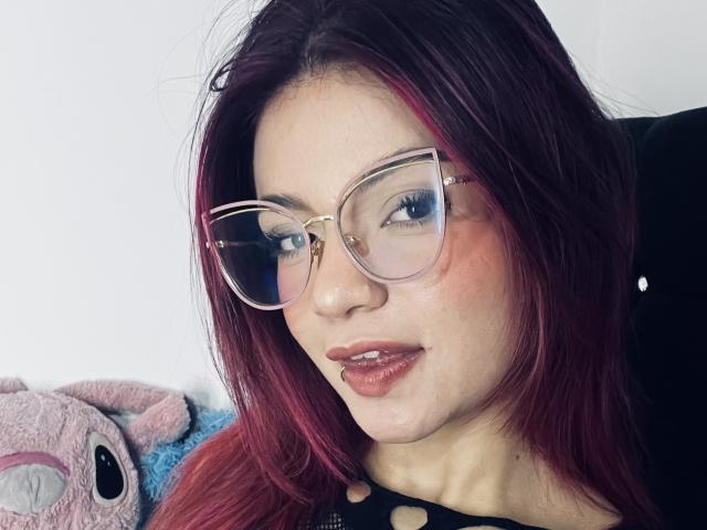 Close-up van GiaSweet met bleke huid, rood haar en een bril met heldere glazen, die verleidelijk in de camera kijkt met licht geopende lippen. lippen.

pluche speelgoed aan de linkerkant. Zachte, sensuele uitstraling..