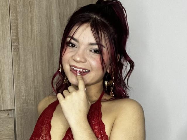 GiaSweet, uma mulher asiática com cabelo ruivo escuro preso em um rabo de cavalo, vestindo um top de renda vermelho com alças, mordendo o dedo. sorriso sedutor, fundo com painéis de madeira, pose sugestiva.