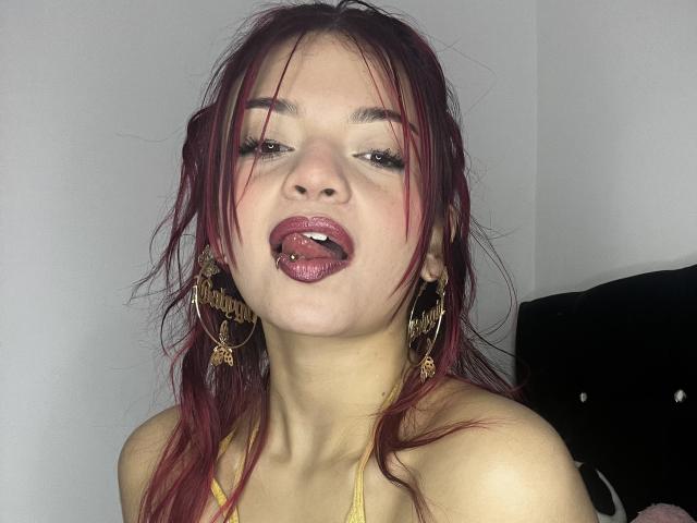 Close-up photo of GiaSweet, Asian woman with wet, red hair, gold hoop earrings, and a yellow halter top. She's licking her lips seductively, eyes half-closed, showing sexual tension.