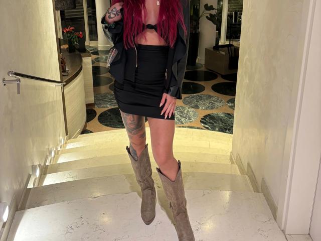 ScarletSins - uma mulher atraente com longos cabelos ruivos, vestindo um top preto sem alças e uma minissaia, botas marrons que chegam até o joelho, pernas tatuadas. Em pé, em uma porta bem iluminada, com o órgão genital mal visível. Pose sensual e provocante.