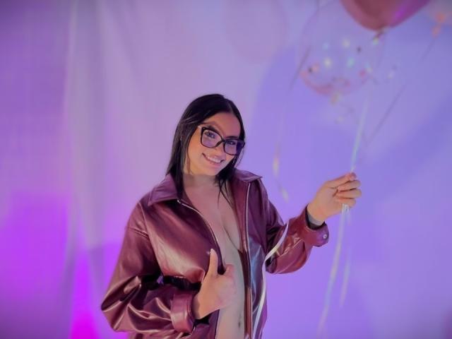 MiiaPalmer mit Brille, lächelnd, die an einem Luftballonseil zieht, trägt eine offene Lederjacke, in den Farben Lila und Blau. Beleuchtung, schwarzes Haar, leicht mollig, sexy, selbstbewusst, lässige Pose, modern, verspielte Atmosphäre..