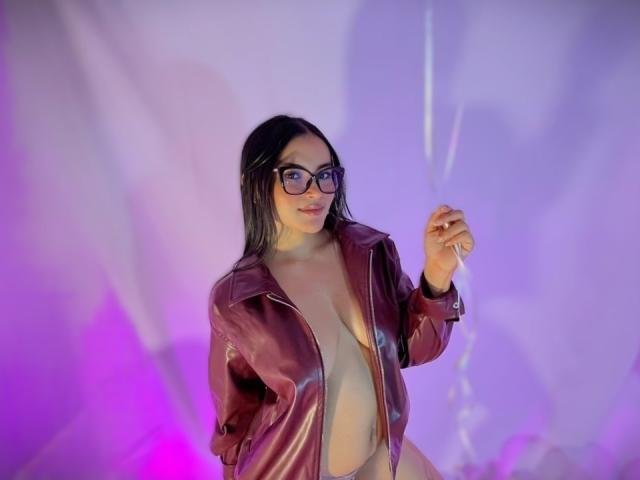 Miia Palmer si erge con sicurezza in una stanza scarsamente illuminata, con luci al neon viola e blu. Indossa una giacca di pelle rossa lucida, aperta per mostrare il suo seno generoso. Lei tiene un bicchiere di vetro trasparente nella mano destra, indossando occhiali con montatura nera..