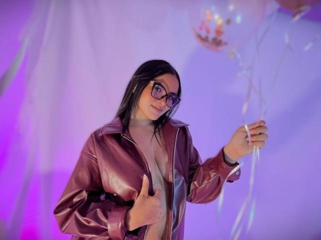 MiiaPalmer, vestida con una chaqueta de cuero roja, está de pie con el pulgar en alto, mostrando su pecho. Ella tiene el pelo negro, usa gafas y tiene una expresión seductora. Luces de neón moradas y azules iluminan el fondo..