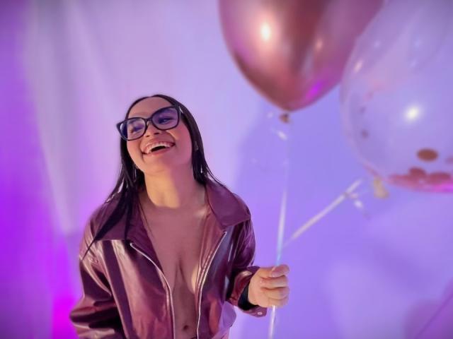 MiiaPalmer, a woman with long black hair and glasses, smiles while holding pink and clear balloons. She wears an open, shiny leather jacket revealing cleavage. Purple and blue lights illuminate her.