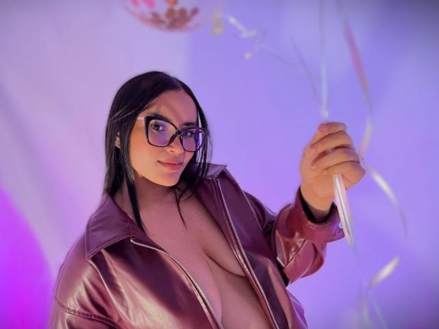 Fotografia de MiiaPalmer, uma mulher curvilínea com seios grandes, vestindo uma jaqueta de couro marrom com o zíper parcialmente aberto, segurando uma varinha brilhante, com luzes roxas e azuis ao fundo. Ela está olhando sedutoramente para a câmera..