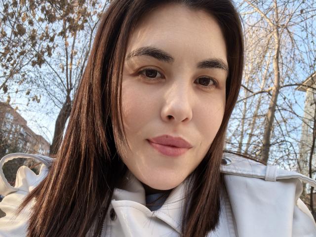 Close-up photo of ErikaGloss with long dark brown hair, fair skin, and brown eyes. She wears a white coat, looking directly at the camera with a slight smile. Outdoor trees in background. Sexual tension from direct gaze.