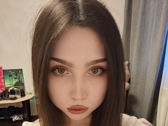 SophiaGlau has pale skin, green eyes, and long brown hair with subtle waves. She has full eyebrows, long eyelashes, and pinkish-brown lipstick. Her expression is neutral and slightly serious.