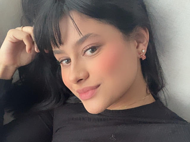 Primer plano de EmmaThomasy, una joven con piel de tono marrón medio, cabello negro y flequillo, sonriendo ligeramente. Ella lleva una blusa negra, pequeños aretes rojos, y está tocándose el pelo. Iluminación suave.