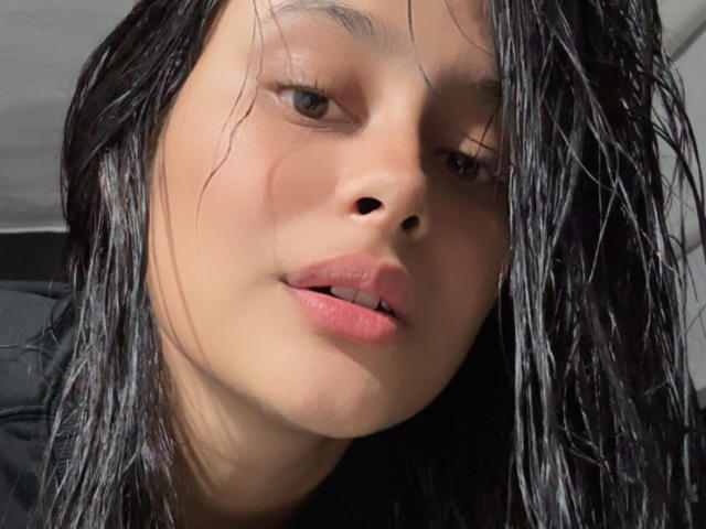 Close-up photo of EmmaThomasy, face filling the frame, wet black hair, slightly parted pink lips, shadowed right eye, light skin, direct gaze, intense expression, subtle light source from upper left.