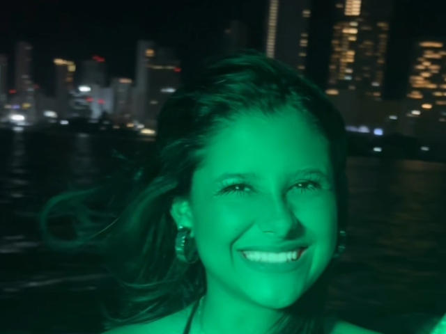 Close-up of EmmaThomasy with green-illuminated face, smiling widely. Dark cityscape with lit skyscrapers in the background. Her hair blows slightly, wearing earrings. Urban night setting, vibrant green light.