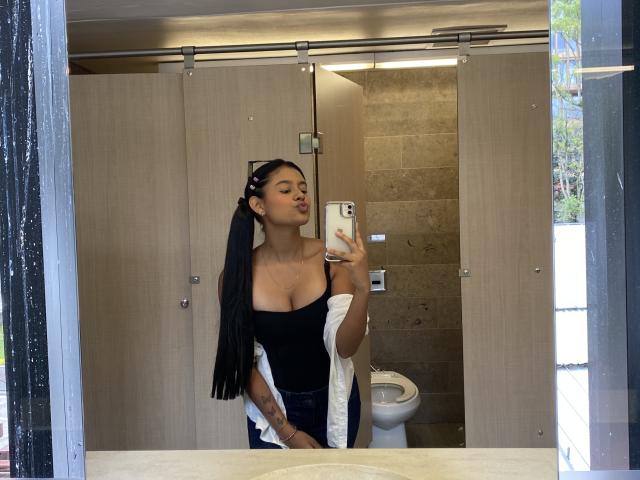 Selfie von EmmaThomasy vor einem Spiegel, sie trägt ein enges schwarzes Kleid mit tiefem Ausschnitt, ein schwarzes Tuch auf dem Kopf und hält ein Handy. Hintergrund eines Badezimmers mit Kabinen und einer Toilette. Verdammt sexy Pose..