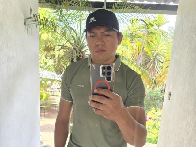 StuardS with a serious face, wearing a black cap and green polo, taking a selfie with an orange ringed phone, standing under a white structure with palm trees.