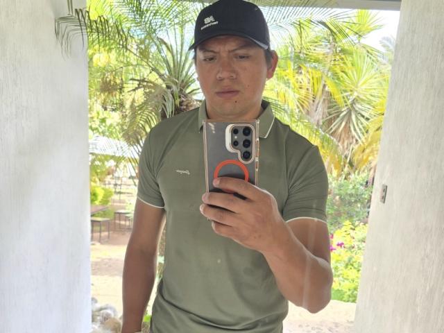 StuardS is a muscular guy with tan skin, wearing a black cap, green polo, and holding a phone with an orange ring, taking a selfie.