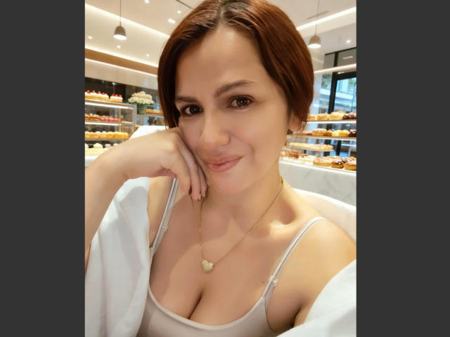 Close-up of CameronViviane in a brightly lit bakery, leaning on her cheek with a playful smile. She has short brown hair, a low-cut beige top, and a gold necklace. Fucking hot.