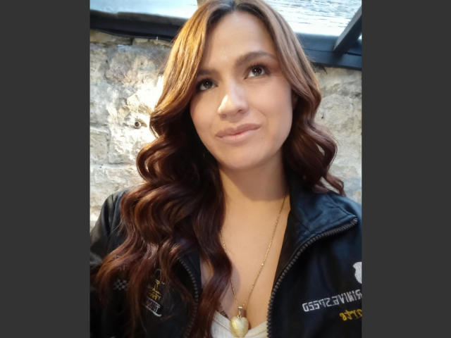 Nahaufnahme von CameronViviane, einer Frau mit langen, welligen braunen Haaren, die eine schwarze Jacke und eine goldene Halskette trägt und lächelt. subtil.
das Licht scheint auf ihr Gesicht und erzeugt einen leichten Schatten an der strukturierten Wand..