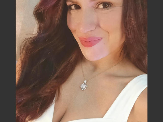 Fotografía de primer plano de CameronViviane, una mujer blanca con cabello castaño largo y ondulado, que lleva una blusa blanca y un collar delicado. con un colgante con forma de flor. Sus labios son rosados y ligeramente entreabiertos. Está mirando directamente a la cámara con una sonrisa sutil. Su escote es visible..