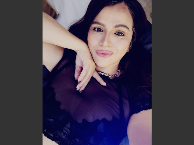 Fotografía de Cameron Viviane, acostada en una cama, vistiendo una blusa negra transparente que revela sus grandes senos. Ella tiene el pelo oscuro, un piercing en la nariz y una sonrisa segura. Su mano descansa sobre su pecho..