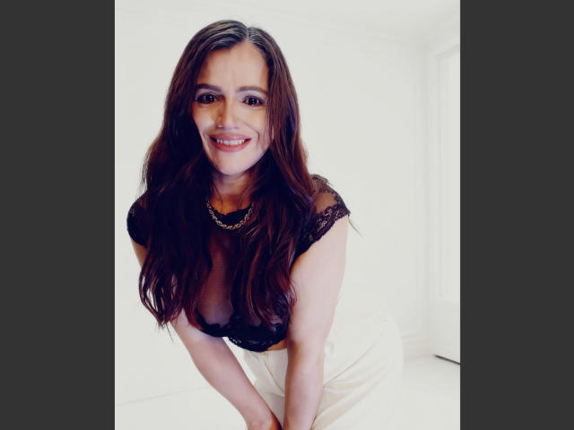 Photograph of a woman, CameronViviane, with long dark brown hair, smiling directly at the camera. She's wearing a black lace top with a deep neckline, sitting on a white couch. Fucking hot.