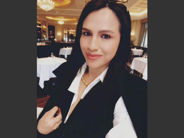Fotografía de CameronViviane, una mujer con cabello largo y negro, que lleva un chaleco negro sobre una camisa blanca, un collar dorado. Sentado en un restaurante con poca luz, con candelabros en el fondo. Su mano está cerca de su pecho, irradiando una confianza sutil y atractiva..