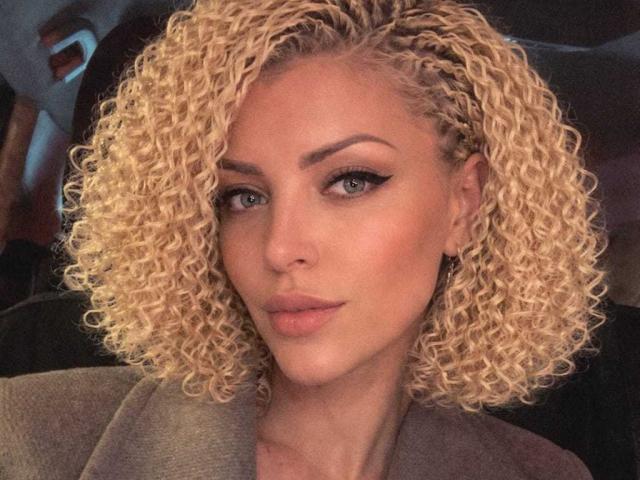 Close-up of Axinia with shoulder-length blonde curly hair, blue eyes, and full lips. She's looking directly at the camera with a slight smile. She's wearing a grey coat. Fucking hot.