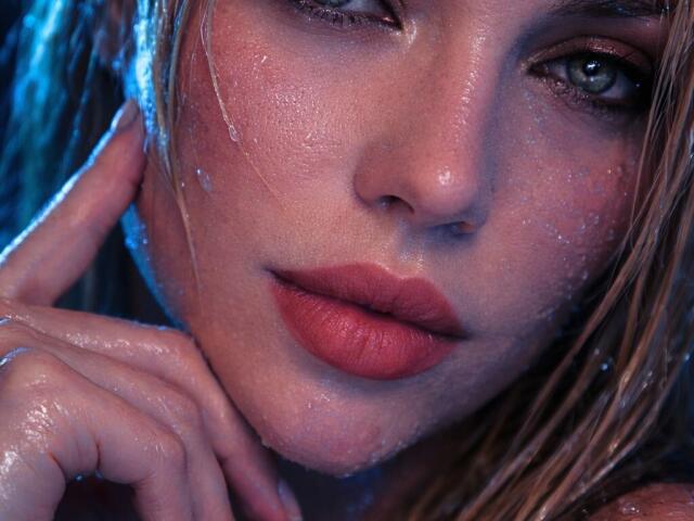 Close-up van Axinia's gezicht, nat en glanzend, met rode lippen die lichtjes open staan. Haar hand raakt haar wang aan, blauw licht accentueert haar blauwe ogen. De huid is glad, wat een gevoel van seksuele spanning oproept..