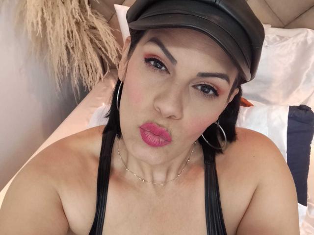 Close-up of LizaRussell, wearing a black cap, hoop earrings, and a black halter top, pouting with bright pink lipstick. Her eyebrows are thick and dark, skin is fair. Background is blurred.