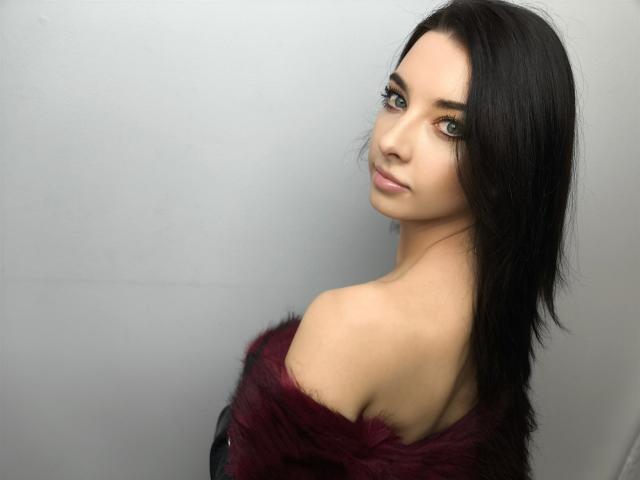 Fotografía de una DominaLuna con cabello negro largo, mirando por encima del hombro, vestida con una prenda de piel burdeos oscuro que se lleva fuera del hombro. piel clara, mirada intensa, ligera sonrisa, luz suave, fondo gris..