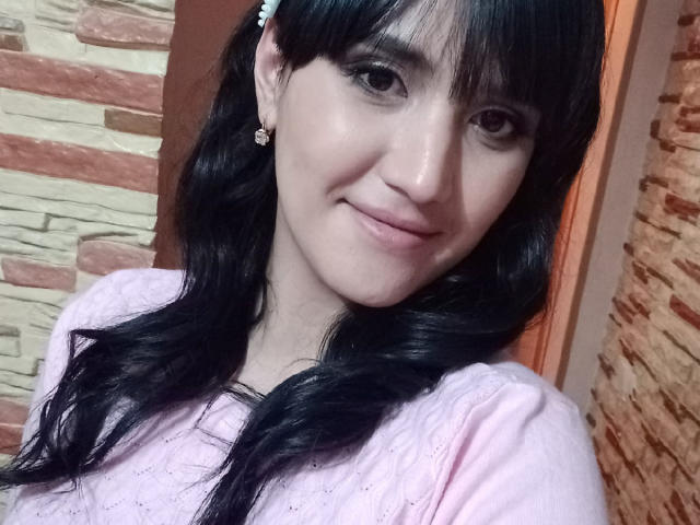 Selfie em close-up de MilanaRawsx, uma mulher de pele clara com cabelo preto e franja, vestindo uma blusa branca, sorrindo discretamente. com uma parede de tijolos e pedras ao fundo. Muito atraente..