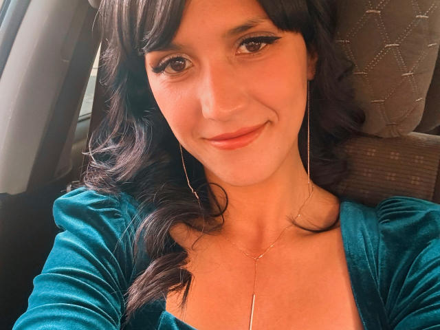 Close-up de MilanaRawsx, sorrindo com cabelos castanhos ondulados, vestindo uma blusa verde-água e um colar dourado, sentada em um carro. A luz do sol realça seu rosto, criando uma aparência calorosa e convidativa..