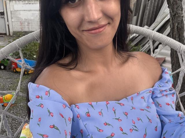 Close-up photo of MilanaRawsx, light-skinned woman with long black hair, smiling, wearing an off-shoulder blue top with red floral patterns, sitting on a white chair, outdoor setting. Visible toy truck.