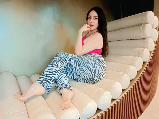 Maby Legrand, mit langen schwarzen Haaren, trägt ein pinkes Oberteil und eine Hose mit Zebra-Muster und entspannt auf einem weißen, gebogenen Sofa, die Füße sitzend auf dem Sitz, verführerisch in die Kamera blickend. Helle Beleuchtung.