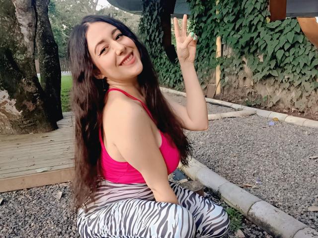 Maby Legrand, con lunghi capelli neri, una canotta rossa e pantaloni a righe zebrate, seduta sulla ghiaia, sorridente e che fa un gesto di pace. Segno, albero e muro ricoperto di edera sullo sfondo. Molto attraente..