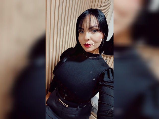 Photograph of JessicaFiore with dark straight hair, red lipstick, and black outfit with puffed sleeves. Blurred figure in foreground. JessicaFiore's confident gaze, slight smirk, and curvy figure. Sexual tension.