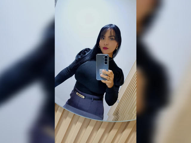 Mirror selfie of JessicaFiore, with straight black hair and red lipstick, holding a phone, wearing a black turtleneck and dark skirt, standing against a white wall, wood-patterned floor. Fucking hot.