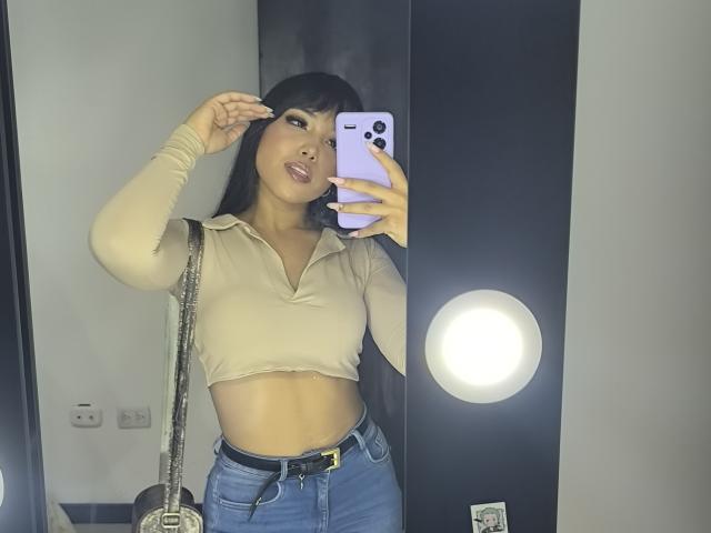 Heyly, taking a mirror selfie, wearing a tight beige crop top, showing a flat stomach, and high-waisted jeans, holding a white phone, with a round light behind her. Fucking sexy.