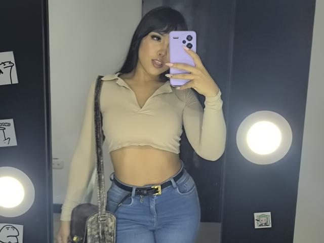 Heyly, com cabelo preto, vestindo uma blusa cropped bege e jeans azuis, tirando uma selfie no espelho com um iPhone. Ela está fazendo beicinho com a língua de fora, usando um cinto preto. Luzes brilhantes..
