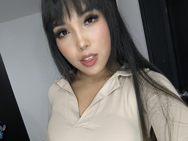 Primo piano di Heyly, una donna asiatica attraente con lunghi capelli neri e frangia, che indossa una camicia bianca con bottoni. Ha labbra rosa lucide e occhi seducenti. La sua espressione è maliziosa..