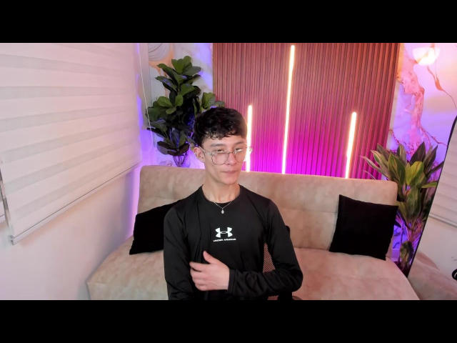 Photograph of PatrickHolland, standing in front of a beige couch with black pillows. He's wearing a black long-sleeve shirt, glasses, and a thumbs-up gesture. Neon lights in the background.