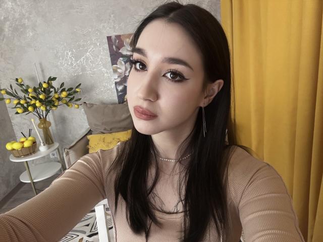 Foto van Alyssa Graham met lang zwart haar, die een beige geribbelde top draagt, grote oorringen en subtiele make-up. Geel gordijn op de achtergrond, gele bloemen en citroenen op de tafel. Heel aantrekkelijk..