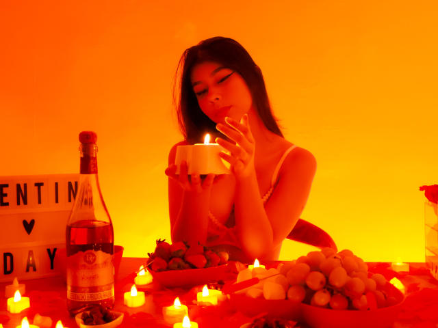 Zoe Winfrey, con i capelli scuri, illuminata da luci rosse, in una posa seducente, mentre sorseggia da un bicchiere, circondata da candele accese, grappoli d'uva e bottiglia, con un cartello che riporta "Festa di San Valentino". Tensione sessuale, luci rosse, espressione sensuale.