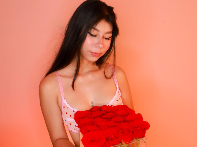 ZoeWinfrey, una donna asiatica attraente con lunghi capelli neri, indossa un reggiseno di pizzo bianco e tiene in mano un grande bouquet di fiori rossi vivaci. rose. Lei sta guardando verso il basso con un sorriso appena accennato.