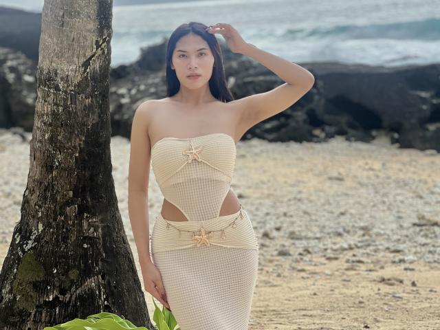 Sofia Cumness está de pie junto a un árbol en una playa, vestida con un vestido sin tirantes, a cuadros y con aberturas, que realza sus curvas. Ella se está tocando la cabeza, con olas del océano al fondo. Increíblemente sexy..