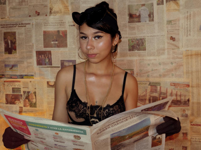 ZoeWinfrey, with black hair in a bun, wearing a lacy black top, gold necklace, and hoop earrings, holding a newspaper, displaying a sultry expression, against a collage of newspaper backgrounds.