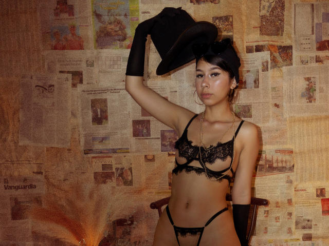 ZoeWinfrey, standing against a textured, collage-like wall, wears black lace lingerie, black gloves, and a black hat. She has an attractive, sexy look with a confident pose, exposing her midriff.